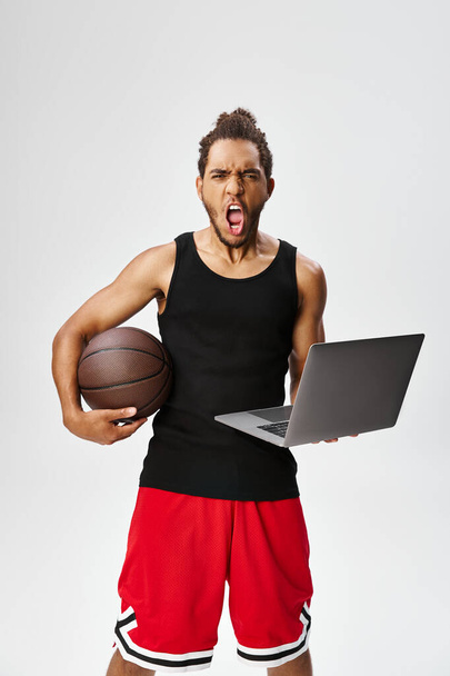 geschokt sportief Afrikaans amerikaanse man met laptop en basketbal kijken naar camera, online wedden - Foto, afbeelding