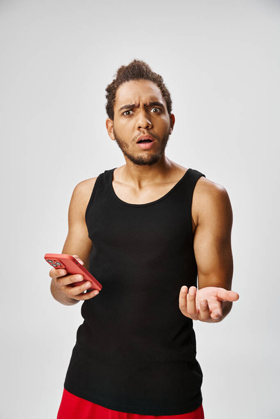 confundido guapo afroamericano hombre sosteniendo el teléfono y mirando a la cámara, apuestas en línea - Foto, imagen