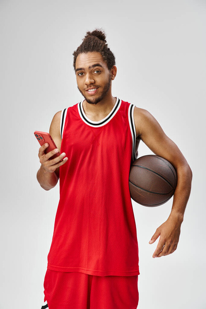 vreugdevolle Afrikaanse Amerikaanse man met basketbal en smartphone en kijkend naar de camera, online wedden - Foto, afbeelding