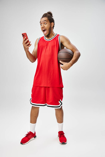 έκπληκτος Αφροαμερικάνος αθλητής κρατώντας μπάσκετ και κοιτάζοντας το τηλέφωνο, online στοιχήματα - Φωτογραφία, εικόνα