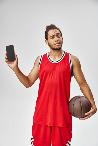 verwarde Afrikaans-Amerikaanse man poseren met telefoon en basketbal en kijken naar camera, online wedden - Foto, afbeelding