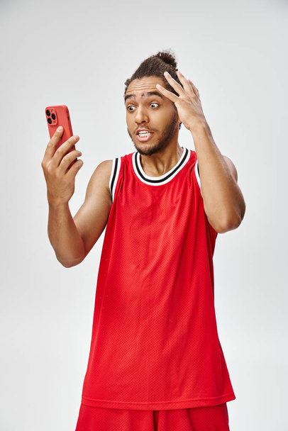 chocado Africano americano desportista olhando para o seu smartphone no fundo cinza, apostas on-line - Foto, Imagem