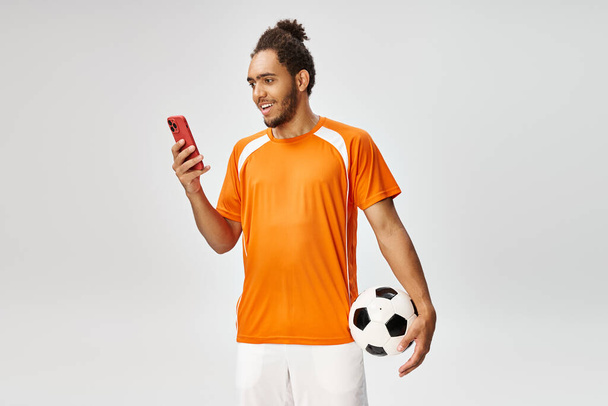 fröhlicher afrikanisch-amerikanischer Mann in Sportbekleidung, der Fußball in der Hand hält und auf das Telefon schaut, Online-Wetten - Foto, Bild