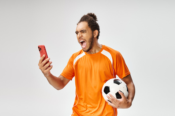 uomo afroamericano emotivo in abbigliamento sportivo in possesso di calcio e guardando il telefono, scommesse online - Foto, immagini