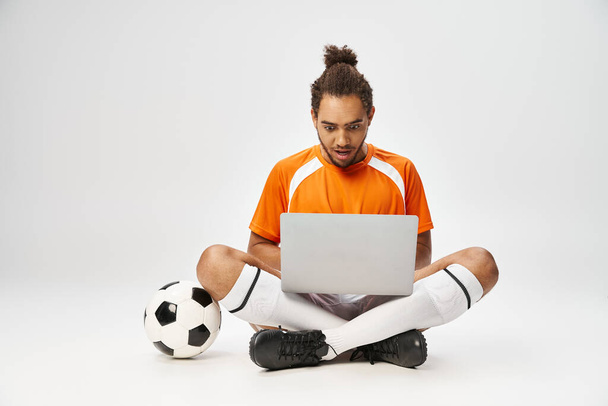 συναισθηματικός Αφροαμερικάνος που κοιτάζει φορητό υπολογιστή κάθεται στο πάτωμα δίπλα στο ποδόσφαιρο, online στοιχήματα - Φωτογραφία, εικόνα