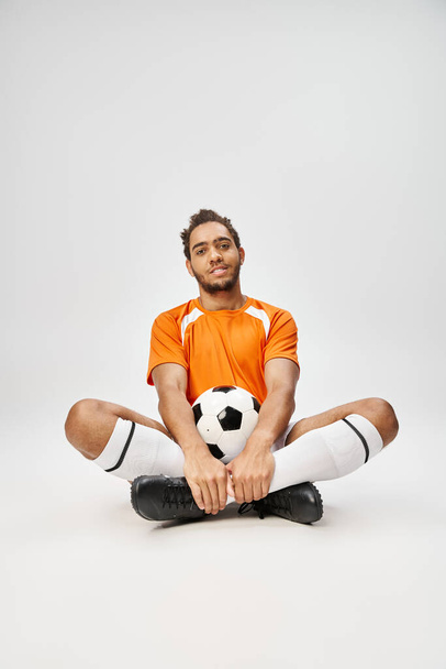 fröhlicher afrikanisch-amerikanischer Mann in gemütlicher Sportbekleidung, der mit Fußball auf dem Boden sitzt und in die Kamera blickt - Foto, Bild