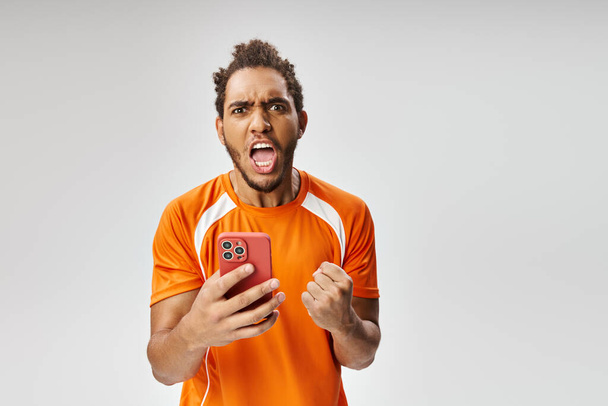 schockierter afrikanisch-amerikanischer Mann in Sportuniform mit Smartphone, der in die Kamera schaut, Online-Wetten - Foto, Bild