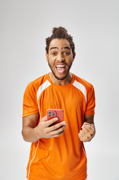 veselý překvapený africký Američan drží smartphone a dívá se na kameru, on-line sázení - Fotografie, Obrázek