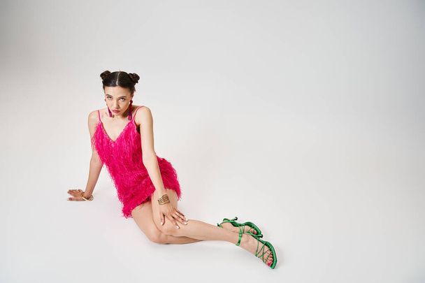 Сияющая дама в розовом платье трогает колено, сидит и смотрит в камеру на сером фоне - Фото, изображение