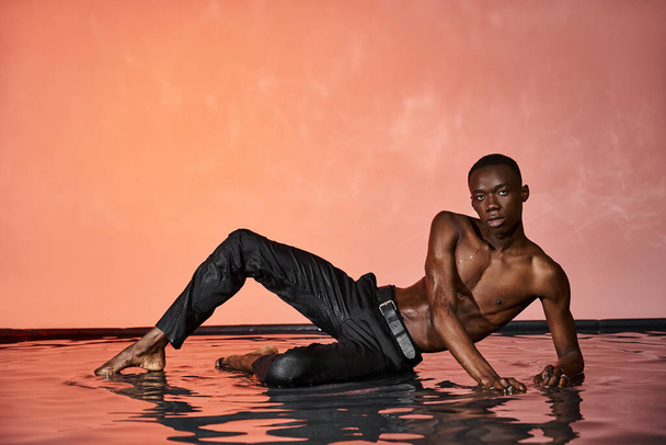beau sans chemise afro-américain en pantalon noir humide posant sur l'eau et regardant la caméra - Photo, image