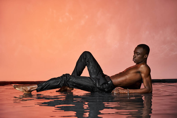 gut aussehende afrikanisch-amerikanische Mann in schwarzen nassen Hosen auf der Wasseroberfläche liegend und wegschauend - Foto, Bild
