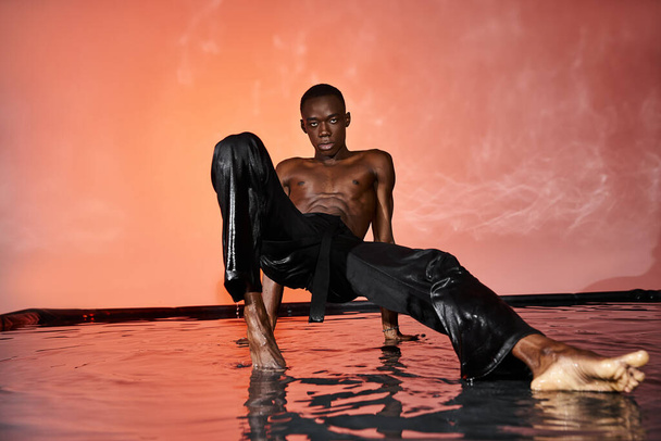 atractivo afroamericano hombre sentado en topless en la superficie del agua y mirando a la cámara, luces - Foto, imagen