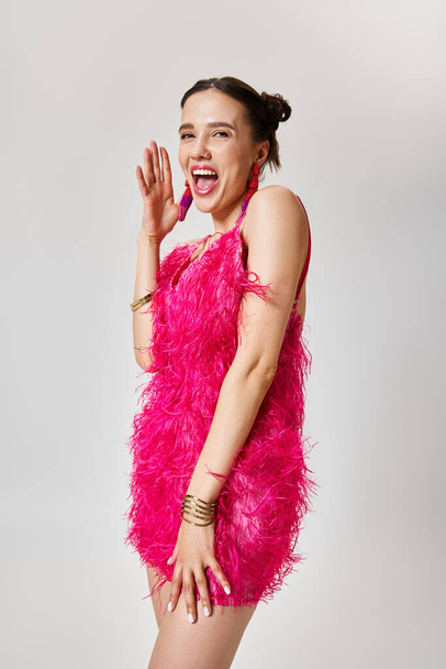 Забавная женщина в стильном розовом платье хихикает, касается щеки, на сером фоне - Фото, изображение