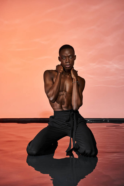 attraente afroamericano uomo in pantaloni bagnati seduto sulla superficie dell'acqua e guardando la fotocamera, luci - Foto, immagini