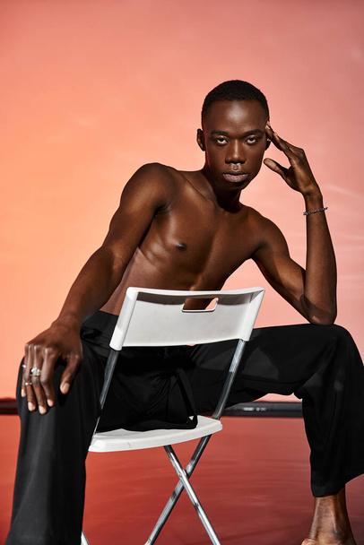pohledný sexy africký Američan sedí bez trička na židli a dívá se na kameru, červená světla - Fotografie, Obrázek