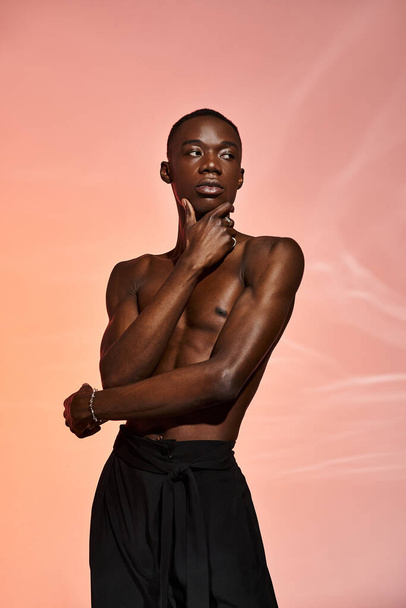 σέξι Αφροαμερικανός που στέκεται γυμνός με μαύρο παντελόνι περιτριγυρισμένος από φώτα και κοιτάζει αλλού. - Φωτογραφία, εικόνα