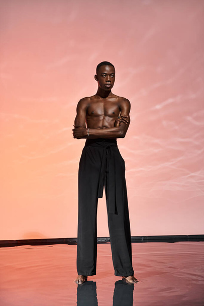sexy africano americano hombre de pie sin camisa en negro pantalones rodeado de luces y mirando hacia otro lado - Foto, Imagen