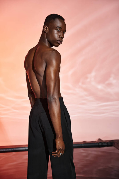 привлекательный африканский мужчина без рубашки стоит и смотрит в камеру в окружении красных фонарей - Фото, изображение