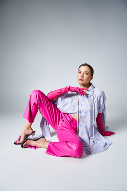 Elegante morena con guantes y pantalones de cuero rosa sentada y agarrada de la mano cerca de la cara - Foto, imagen