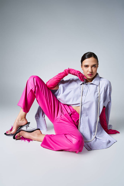 Attractive woman in pink pants and gloves, violet shirt, sitting confidently and touching her chin - Photo, Image