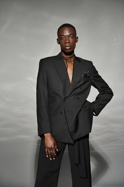 συμμετοχή αφροαμερικανός άνδρας σε ξεκούμπωτο μαύρο κοστούμι ποζάρουν alluringly και κοιτάζοντας την κάμερα - Φωτογραφία, εικόνα