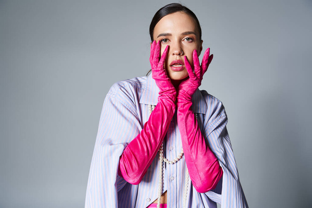 Portret van fashion-forward brunette meisje in roze handschoenen, raakt haar gezicht op grijze achtergrond - Foto, afbeelding