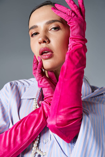 Brunette vrouw met piercing in stijlvolle outfit en roze handschoenen, raakt haar gezicht kijken naar camera - Foto, afbeelding