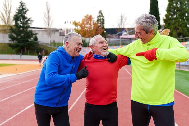 Eine Gruppe fröhlicher älterer Freunde in Sportkleidung teilt ein Lachen und freundliche Gesten nach dem Lauf auf einer Outdoor-Leichtathletikbahn. - Foto, Bild