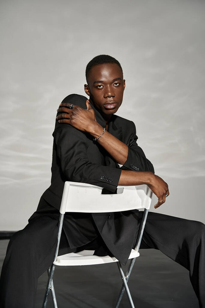 σαγηνευτικό σέξι αφροαμερικανός άνδρας σε κομψό μαύρο κοστούμι κάθεται στην καρέκλα και κοιτάζοντας την κάμερα - Φωτογραφία, εικόνα