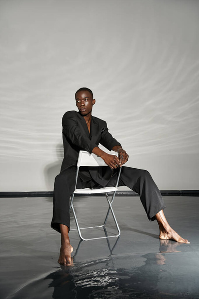 σαγηνευτικό μοντέρνο αφροαμερικανός άνδρας με μαύρο κοστούμι κάθεται και κοιτάζοντας μακριά σε υδαρές φόντο - Φωτογραφία, εικόνα