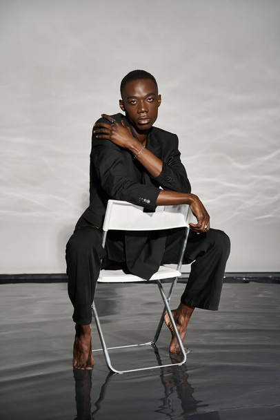 atraente homem americano africano sexy em elegante terno preto sentado na cadeira e olhando para a câmera - Foto, Imagem