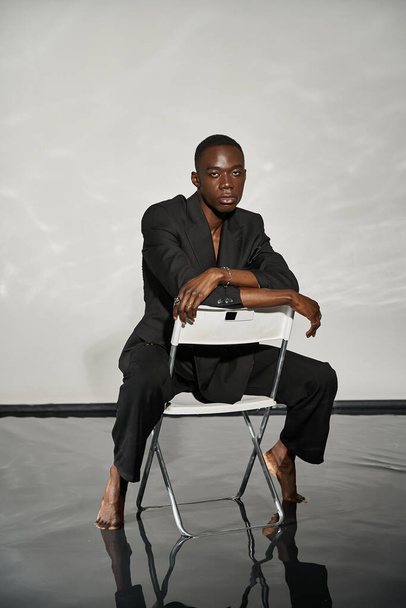 красивий сексуальний афроамериканський чоловік в елегантному чорному костюмі сидить на стільці і дивиться на камеру - Фото, зображення