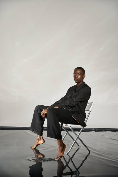 модний сексуальний афроамериканський чоловік в елегантному чорному костюмі сидить на стільці і дивиться на камеру - Фото, зображення