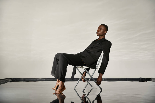 Siyah takım elbiseli şık Afro-Amerikan adam, sulu arka planda kapalı gözlerle sandalyede oturuyor. - Fotoğraf, Görsel