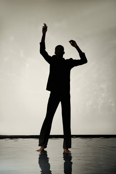 Silhouette eines trendigen afrikanisch-amerikanischen Mannes, der barfuß im Anzug vor grauem, wässrigem Hintergrund im Schatten posiert - Foto, Bild