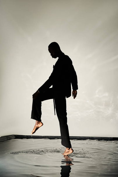 silhouette di affascinante uomo africano americano in posa a piedi nudi in tuta su sfondo acquoso in ombre - Foto, immagini