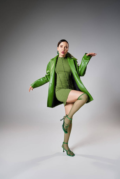 Fashion-forward brunette in green mini dress, leather jacket, knee socks dancing on grey background - Photo, Image