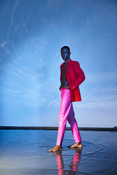 atractivo hombre afroamericano con estilo en pantalones de color rosa y chaqueta roja posando sobre fondo azul acuoso - Foto, Imagen