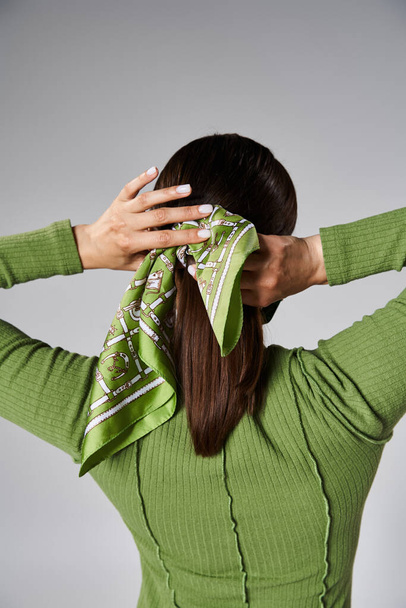 Back view of young brunette woman in total green outfit tying her head scarf on grey background - Photo, Image