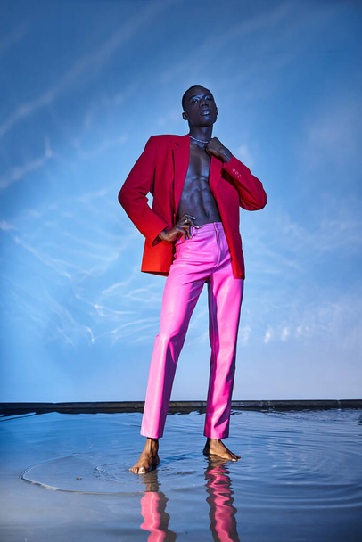ピンクのズボンと赤いブレーザーでスタイリッシュなアフリカ系アメリカ人男性を魅了し,青い水色の背景にポーズ - 写真・画像