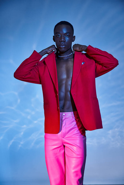 verführerisch stylischer afrikanisch-amerikanischer Mann in pinkfarbener Hose und rotem Blazer posiert vor blauem, wässrigem Hintergrund - Foto, Bild