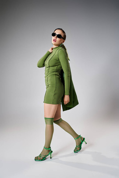 Yeşil elbiseli, güneş gözlüklü şık bir kadın. Omzunda deri ceket taşıyor. - Fotoğraf, Görsel