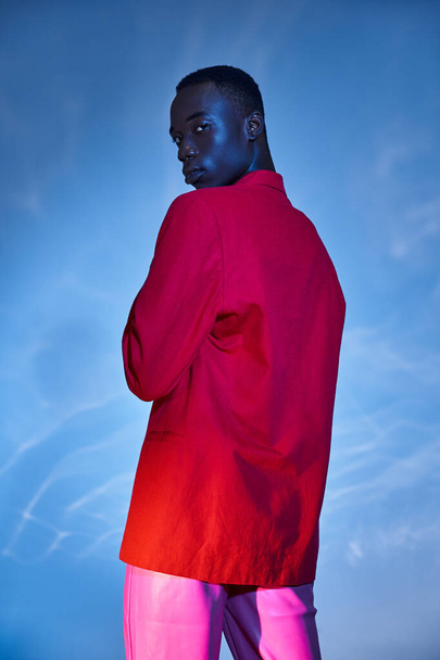 viehättävä tyylikäs afrikkalainen amerikkalainen mies vaaleanpunaisissa housuissa ja punainen bleiseri poseeraa sinisellä vetisellä taustalla - Valokuva, kuva
