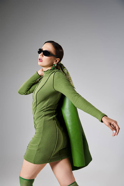 緑の衣装とサングラスのブルネットガールは,彼女の肩に革のジャケットを保持し,ステップを作る - 写真・画像