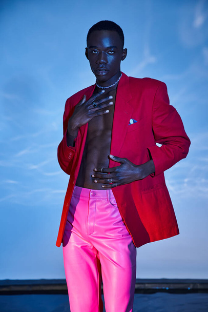 όμορφος κομψός Αφροαμερικάνος με ροζ παντελόνι και κόκκινο σακάκι ποζάροντας σε μπλε υδαρές φόντο - Φωτογραφία, εικόνα