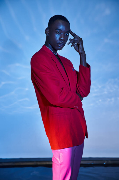 ansprechende stylische afrikanisch-amerikanische Mann in rosa Hose und rotem Blazer posiert auf blauem wässrigem Hintergrund - Foto, Bild