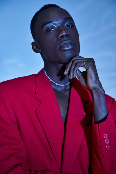 Çekici, Afro-Amerikalı, kırmızı renkli ceketli, mavi sulu arka planda poz veren genç bir adam. - Fotoğraf, Görsel