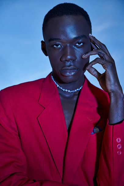 attraktive junge afrikanisch-amerikanische Mann in roten pulsierenden Blazer posiert auf blauem wässrigem Hintergrund - Foto, Bild