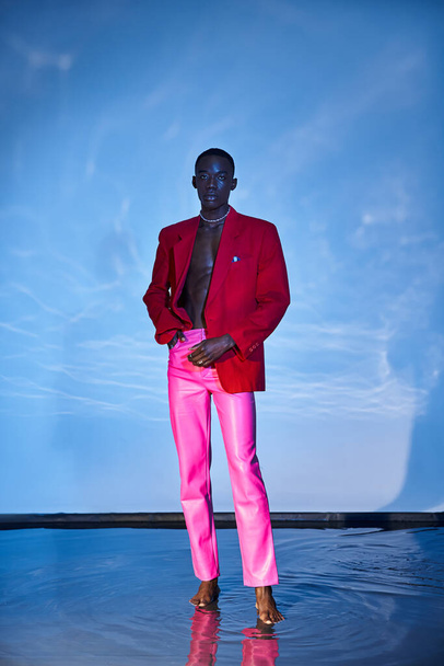 svůdný stylový africký Američan v růžových kalhotách a červené sako pózující na modrém vodnatém pozadí - Fotografie, Obrázek