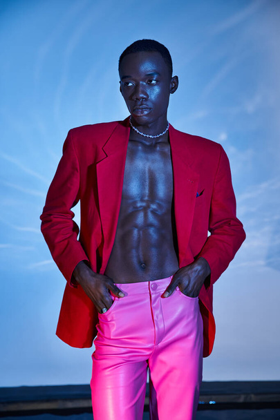 Pembe pantolonlu ve kırmızı ceketli Afro-Amerikan bir adam sulu arka plana bakıyor. - Fotoğraf, Görsel